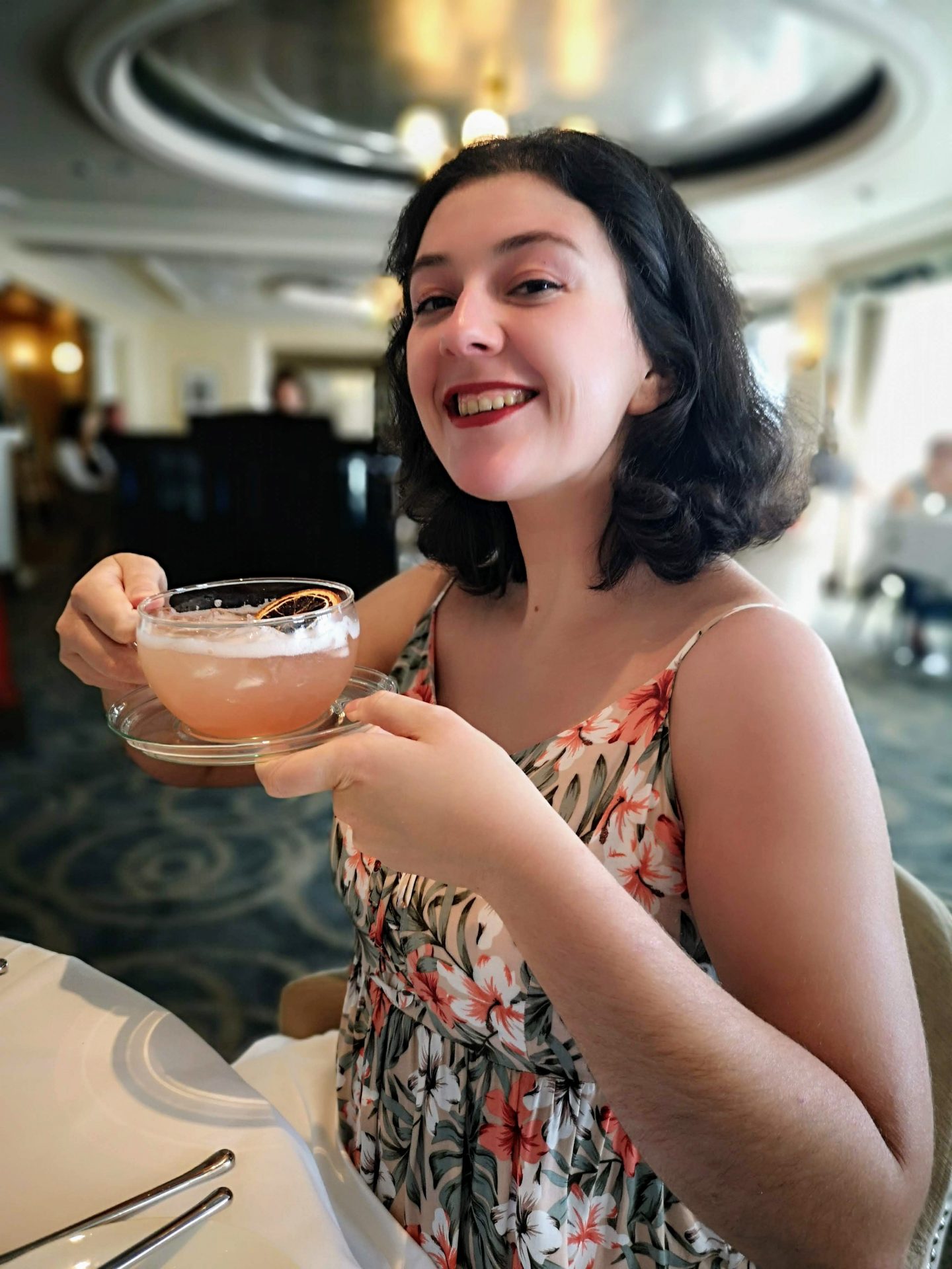 A young women in a floral dress holds a giant glass cup and saucer. The glass cup is full of an amber liquid, not tea but a gin and fruit tea cocktail!