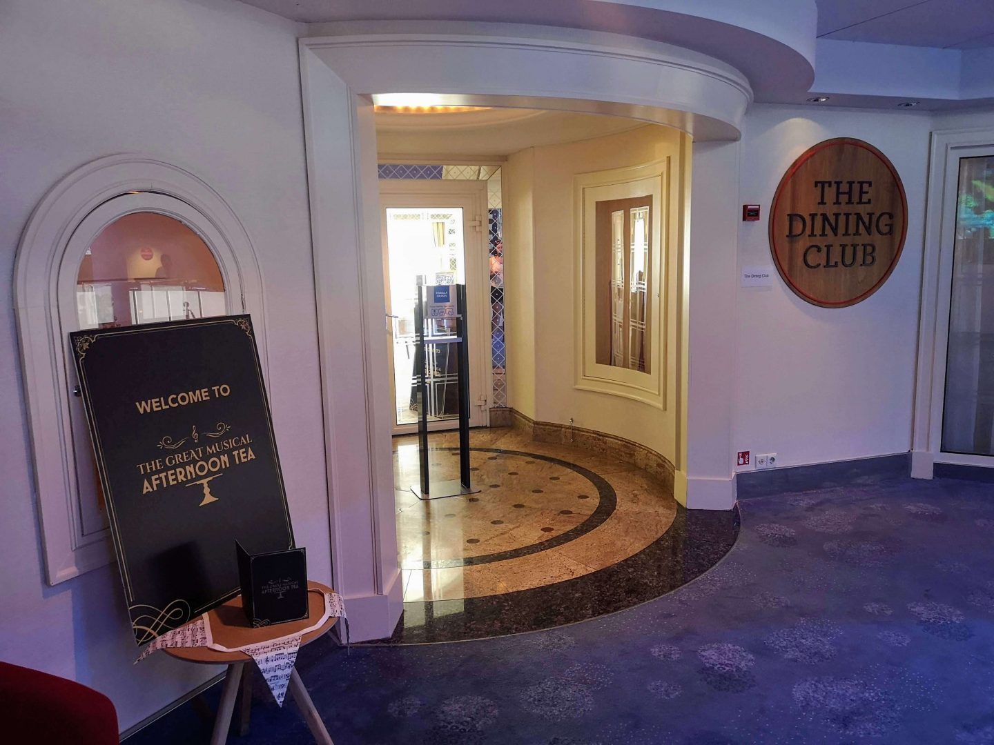 The entrance to Marella Explorer 2's The Dining Club. To the left of the entrance is a table displaying a poster for the Great Musical Afternoon Tea