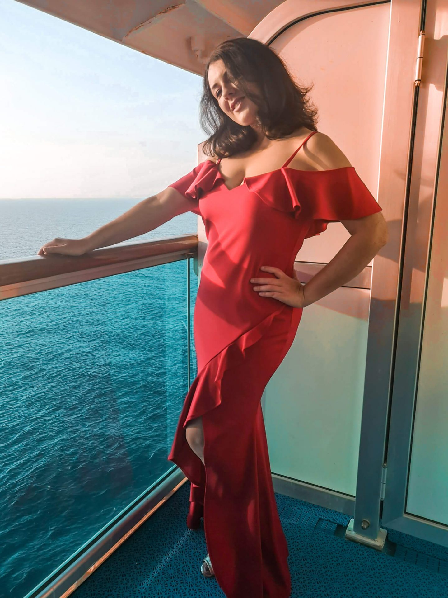 Glammed up on a formal night on a British Isles Cruise - Francesca stands on a ship balcony wearing a red bardot floor length dress with a ruffle up the thigh slit (dress from Quiz Clothing)